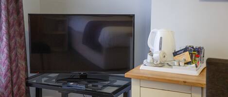 Habitación con televisión de pantalla plana sobre una mesa de vidrio, cafetera y una pequeña alacena con cajones.