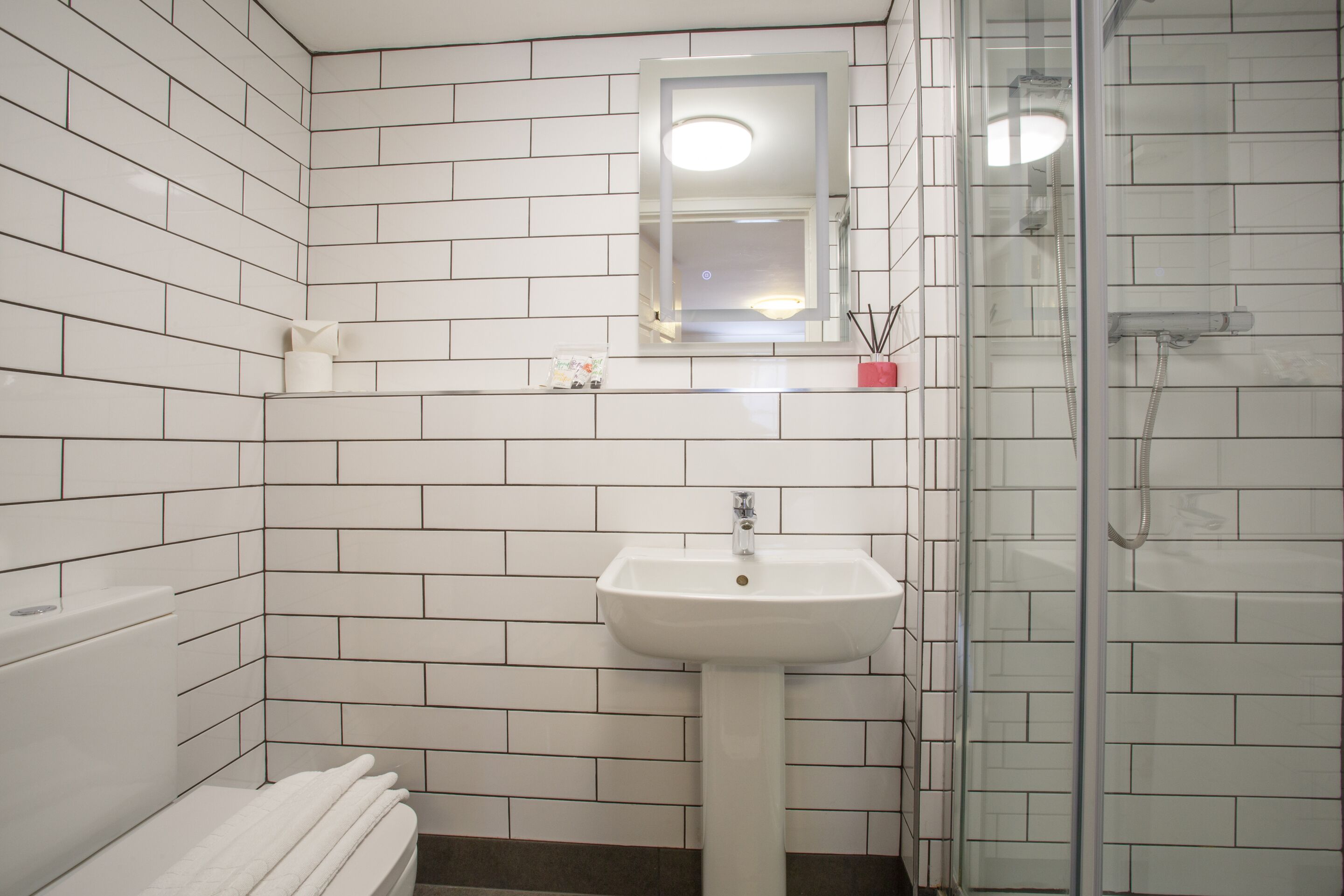 Ein Badezimmer mit weißen Fliesen, einem Waschbecken, einem Spiegel und einer Dusche.