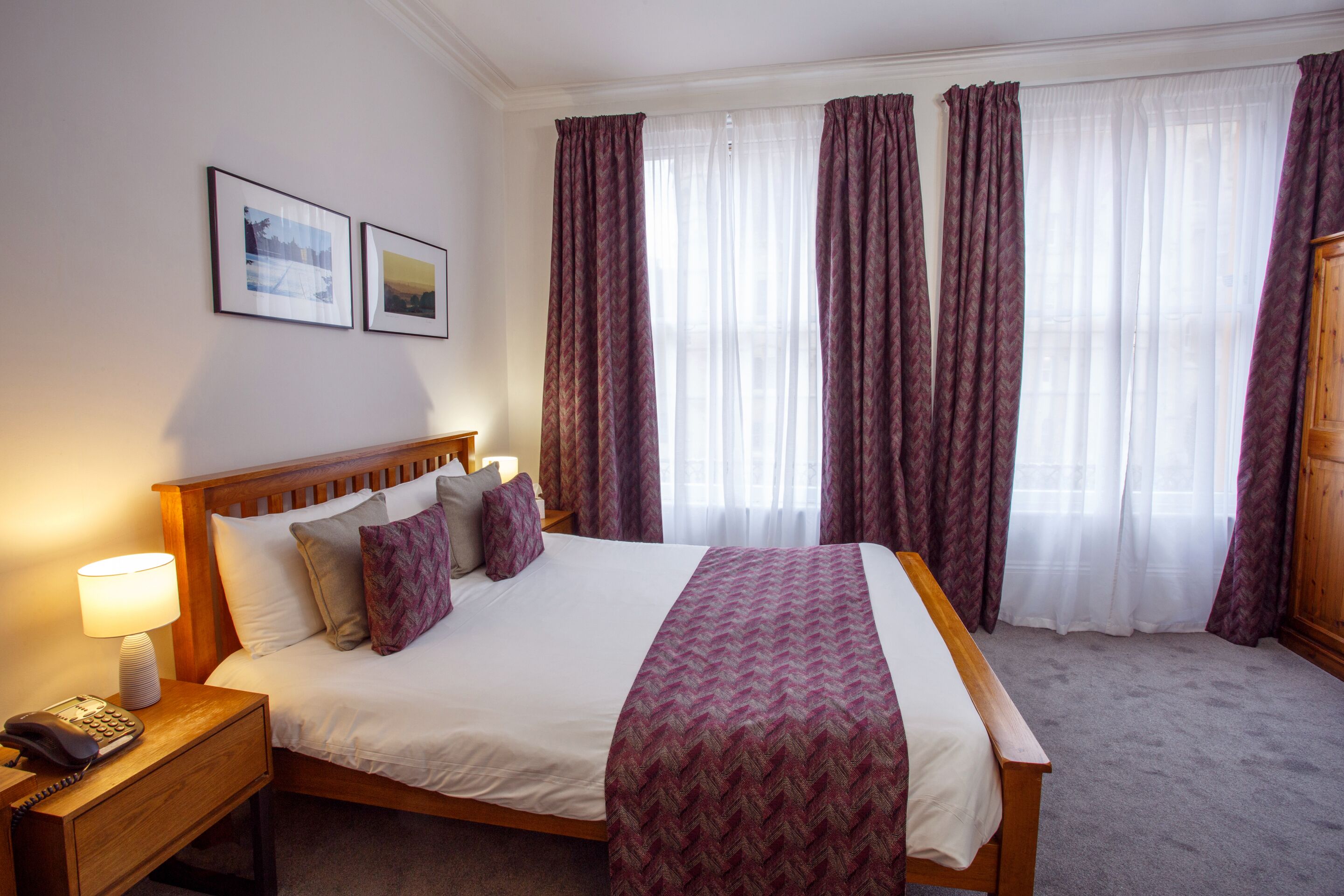 A hotel room with a wooden bed, a nightstand with a lamp and phone, and a large window with curtains.