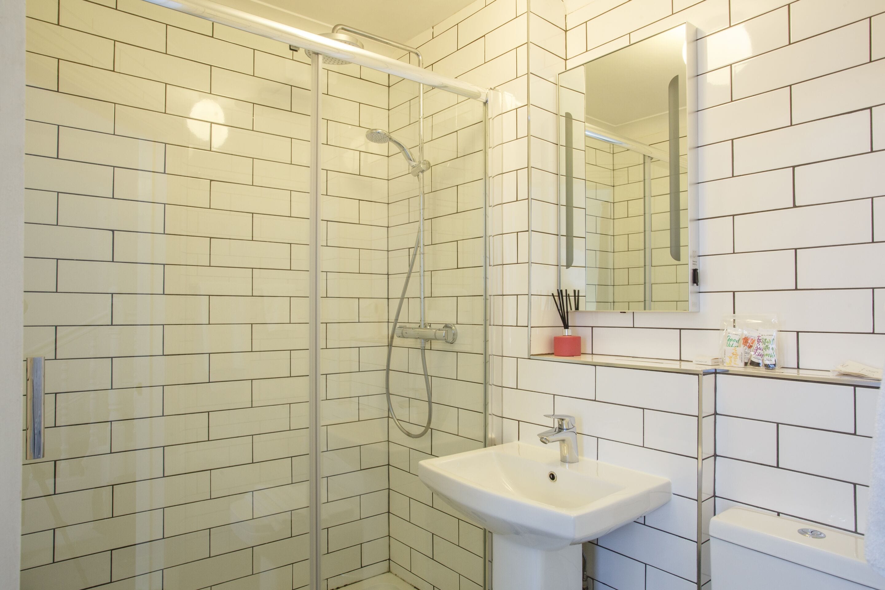 Un baño moderno con azulejos blancos tipo metro, ducha con mampara de vidrio, lavamanos blanco y espejo encima.
