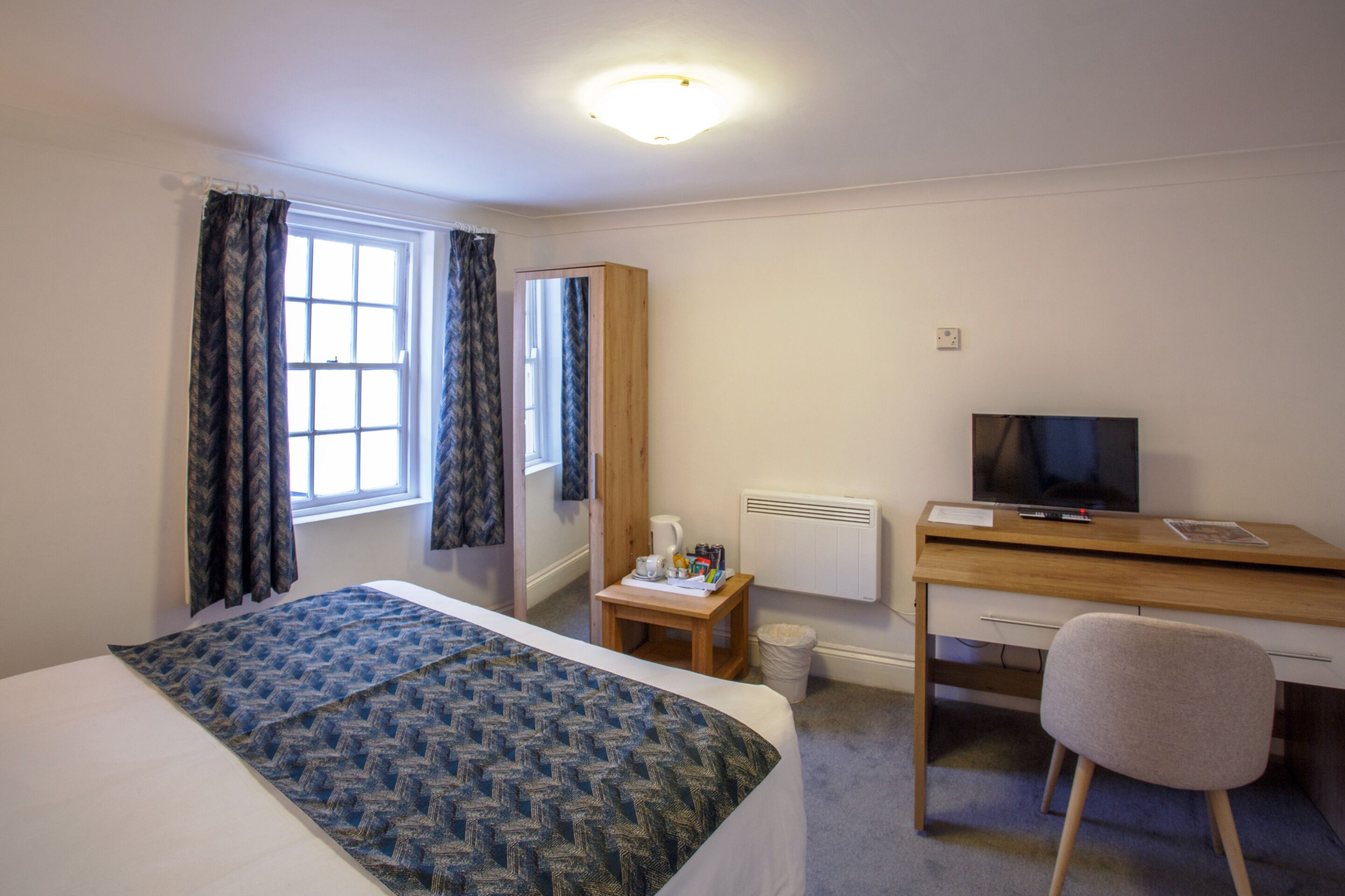 Quarto de hotel com cama, escrivaninha, cadeira, televisão e janela com cortinas.