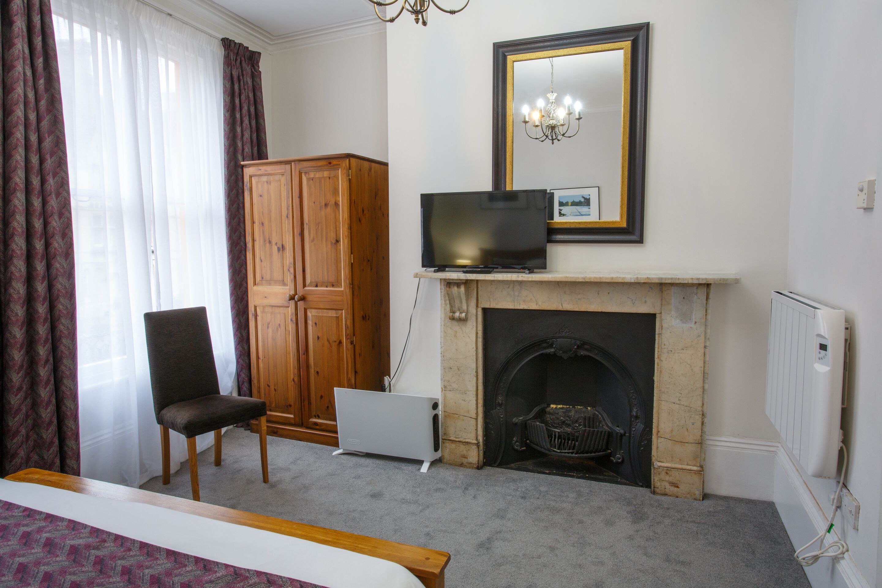 Une chambre à coucher avec un lit, une armoire en bois, un téléviseur posé sur la cheminée, une cheminée et un radiateur.