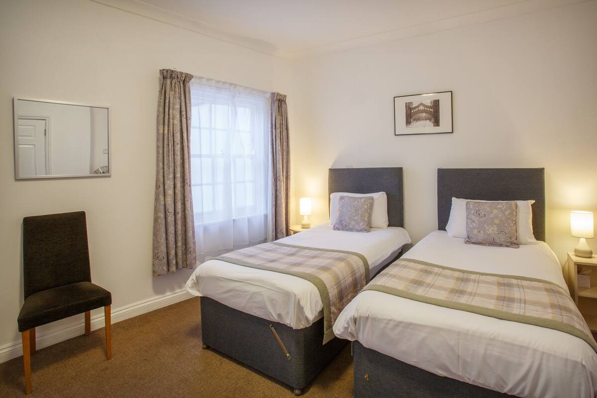 A hotel room with two beds, a chair, a window with curtains, and a framed picture on the wall.