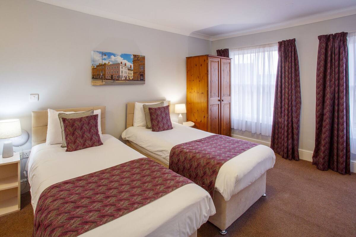 A hotel room with two beds, a wooden wardrobe, a painting on the wall, and a window with curtains.