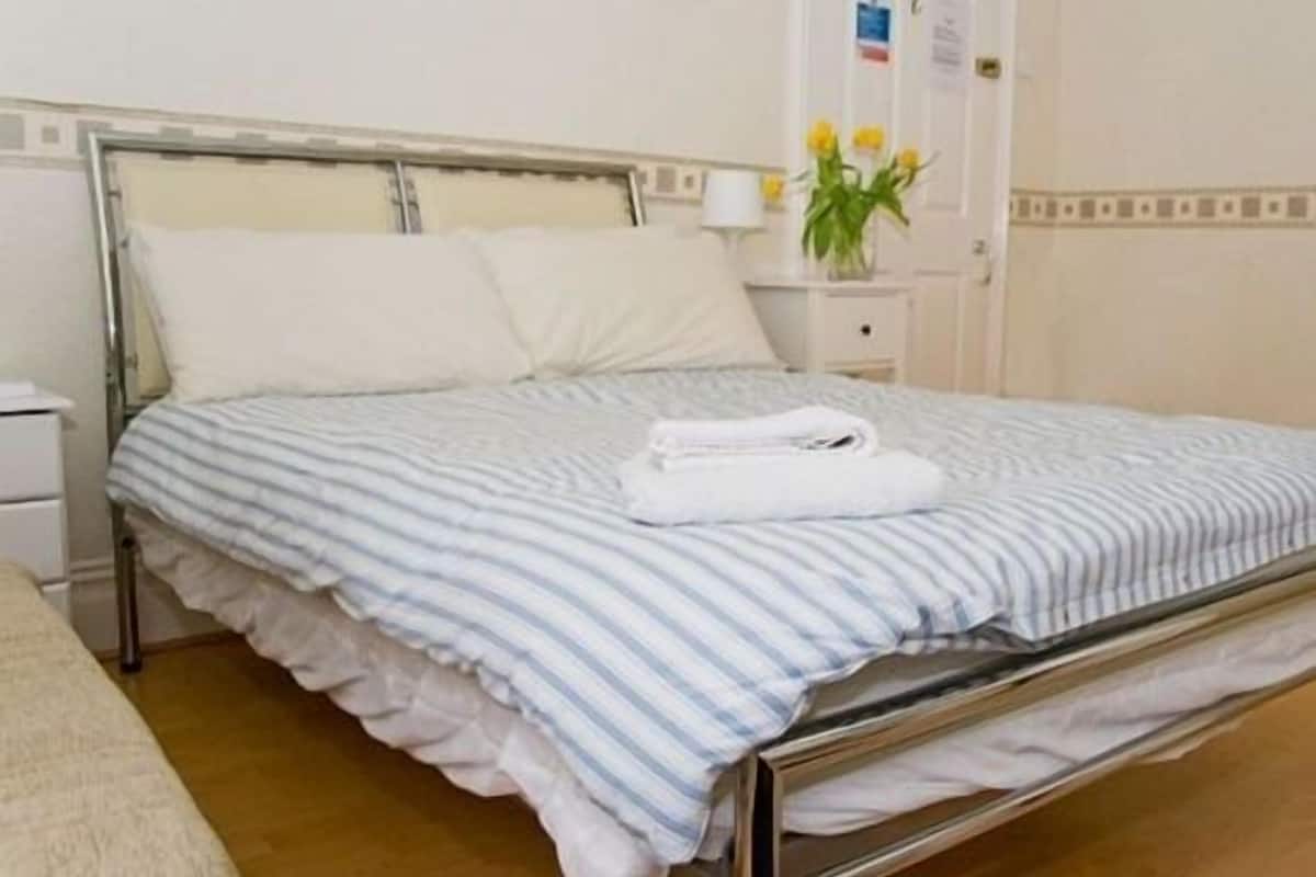 Una cama individual con colcha a rayas y almohadas, una mesita de noche con lámpara, y un florero con flores.