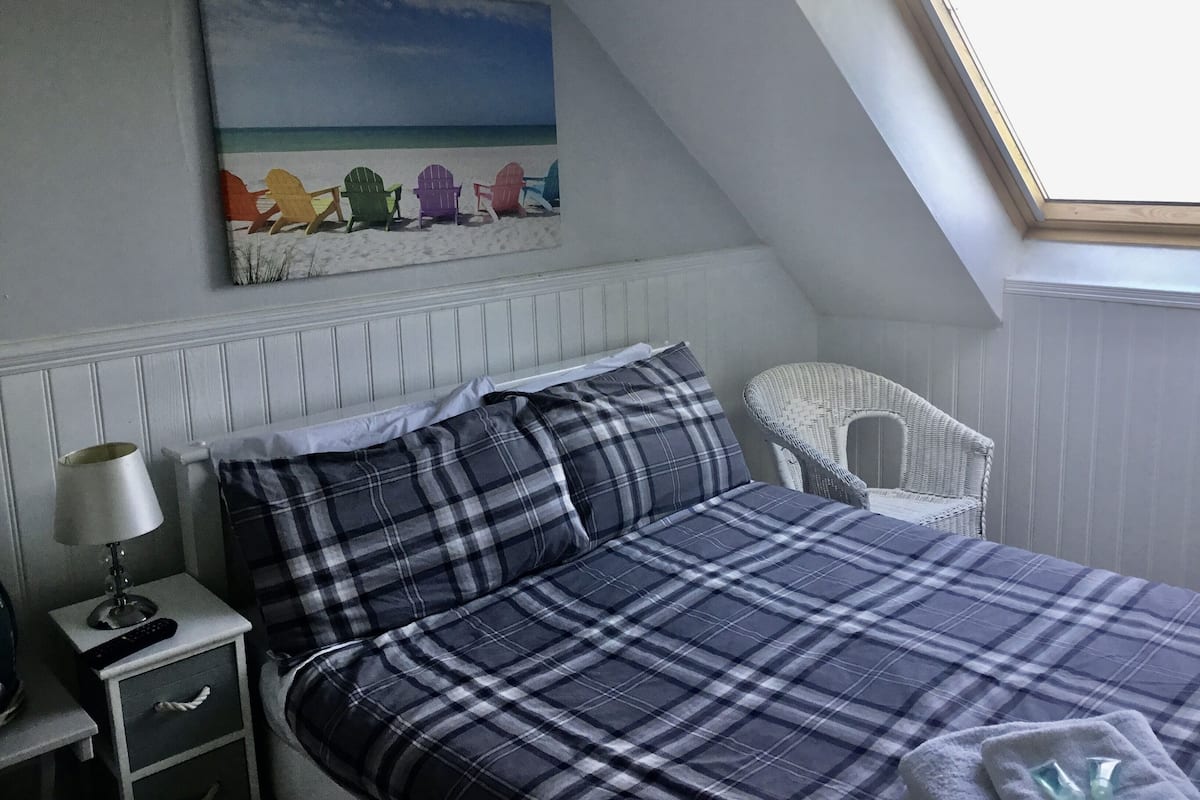 Un dormitorio con techo inclinado, una cama con colcha escocesa, una mesita de noche con lámpara, una silla de mimbre y un cuadro de sillas de playa.