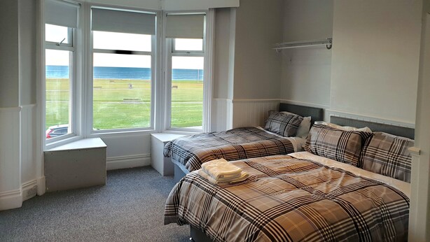 A bedroom with two beds, large windows with a view of a grassy field and the sea, and a white ceiling with recessed lighting. - The Cara Guesthouse (Whitley Bay)
