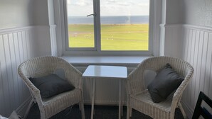 A room with two wicker chairs, a small white table, and a large window offering a view of a grassy field and the sea. - The Cara Guesthouse (Whitley Bay)