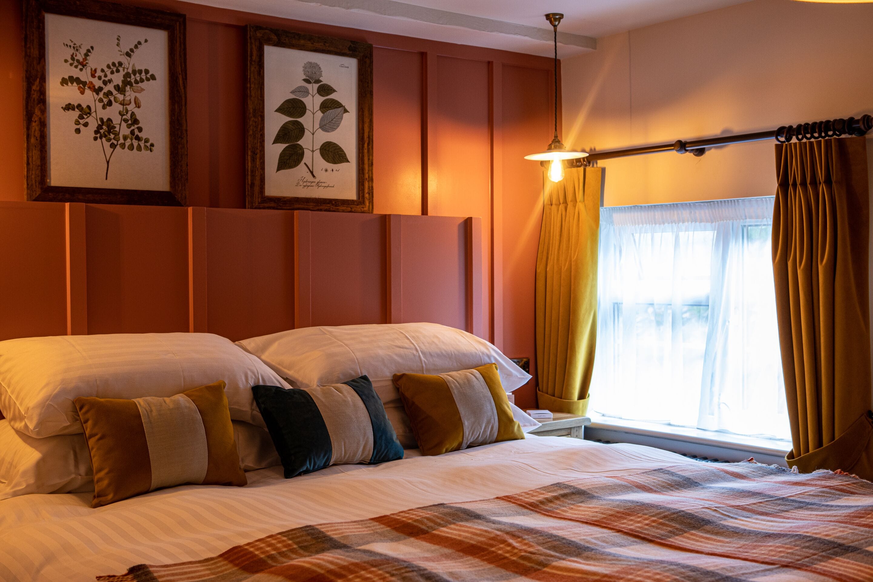 A neatly made bed with multiple pillows, framed botanical prints on the wall, a window with curtains, and a hanging light fixture.