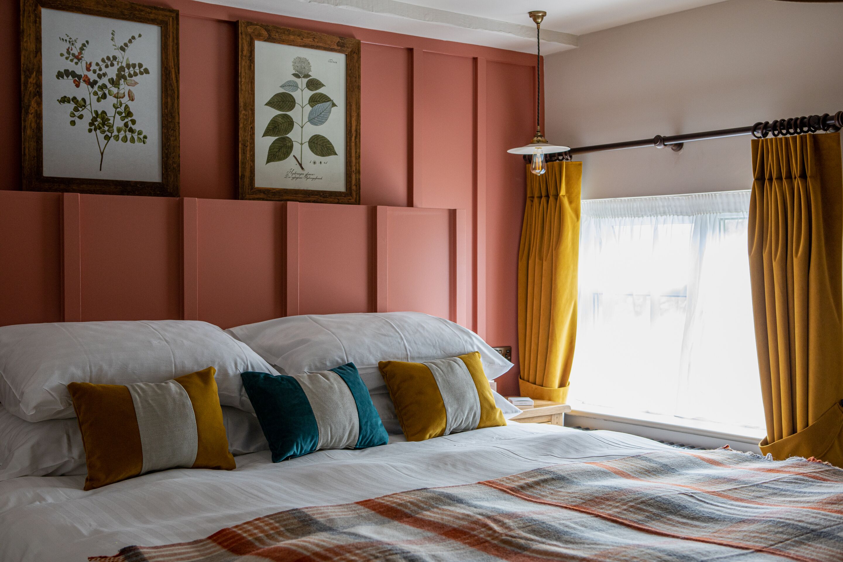 Una cama bien tendida con almohadas blancas y de colores, una colcha estampada e impresiones botánicas enmarcadas en la pared.