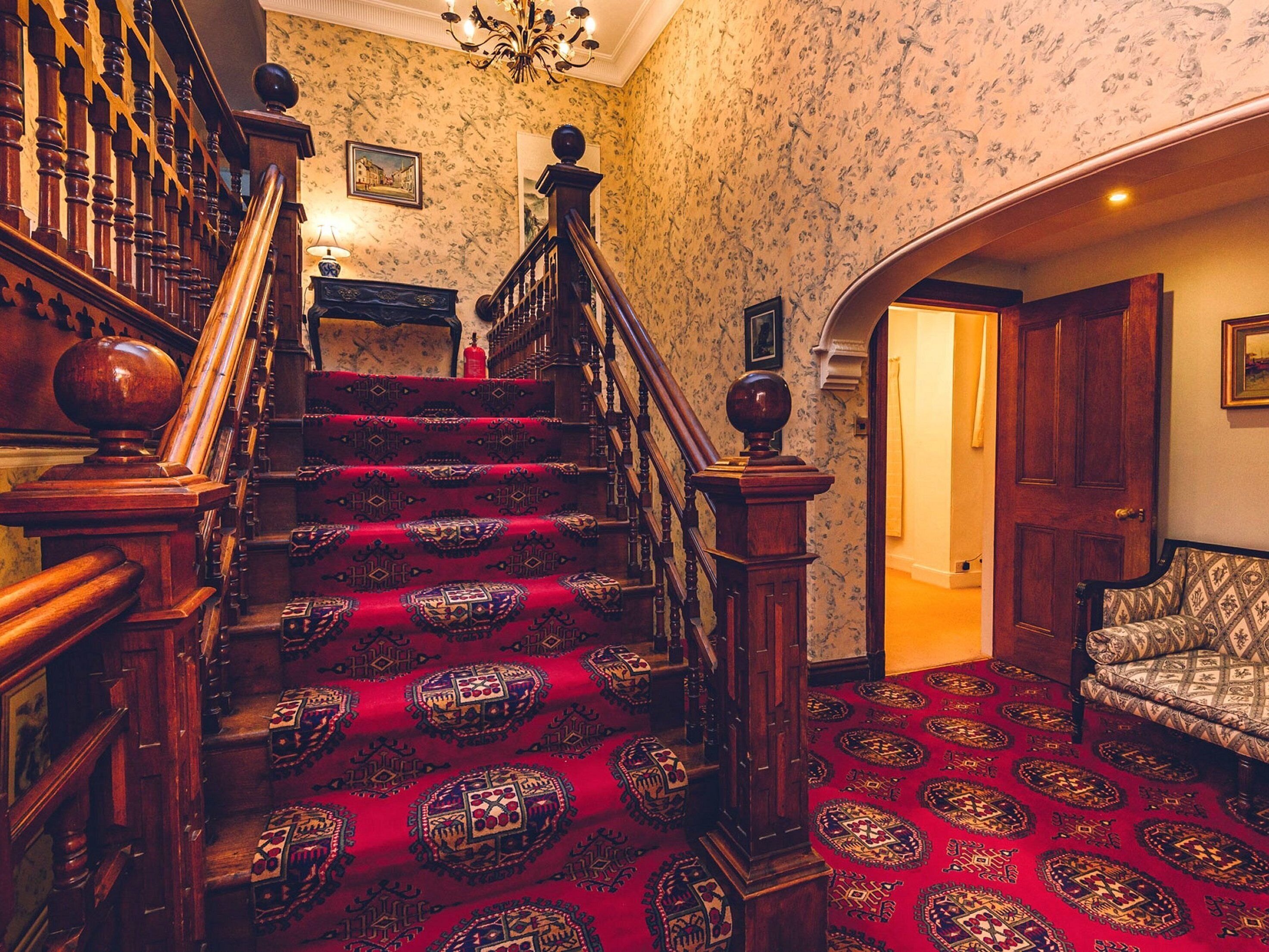 Una imponente escalera con barandales decorados y un suelo cubierto de alfombra roja, que conduce a una habitación con alfombra estampada y una puerta de madera.