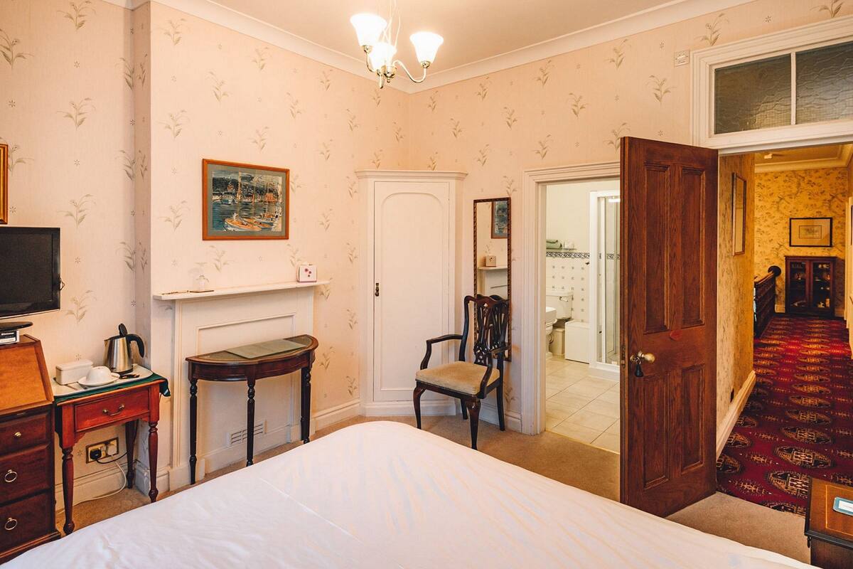 Ein Schlafzimmer mit einem Bett, einem Fernseher, einem kleinen Tisch, einem Stuhl, einem Kamin und einer Tür zu einem anderen Raum.