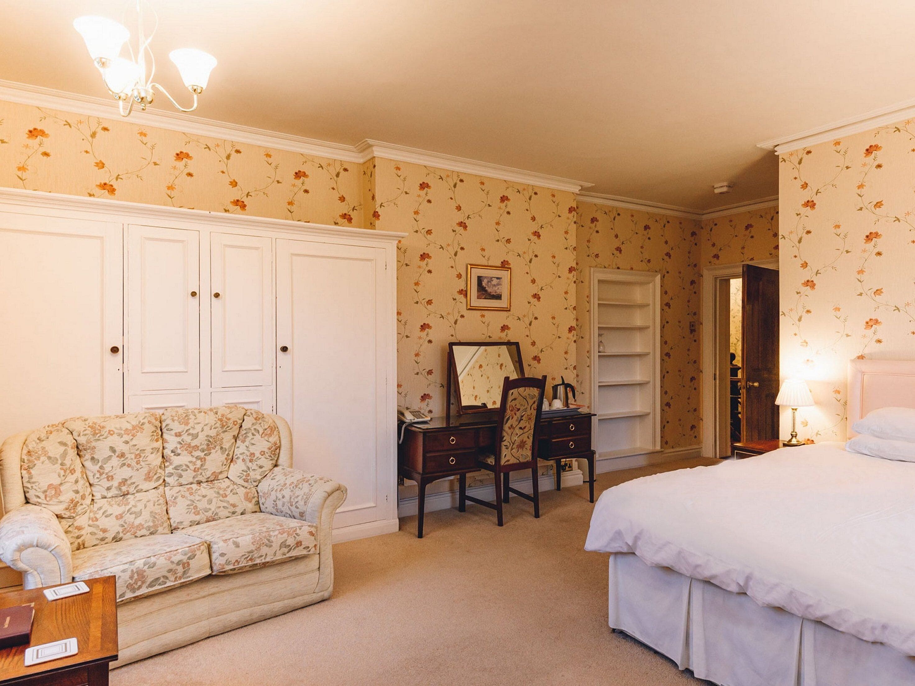 A bedroom with a floral-patterned sofa, a bedside table, a chair, a dresser, a mirror, and a bed.