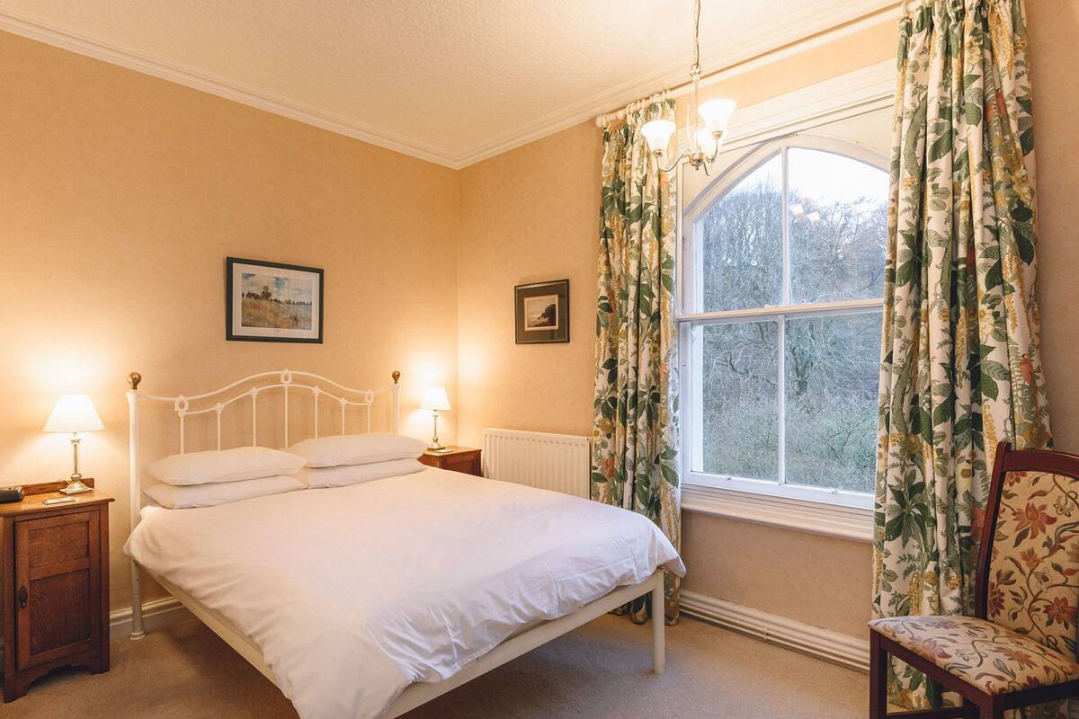Ein Schlafzimmer mit einem Einzelbett, zwei Nachttischen mit Lampen, einem Sessel, einem Fenster mit Vorhängen und zwei gerahmten Bildern an der Wand.