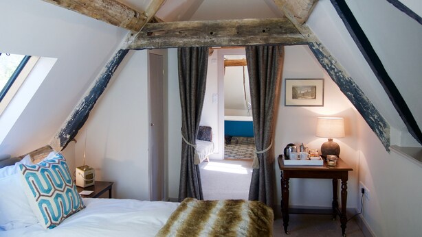 A cozy attic bedroom with exposed wooden beams, a bed with patterned pillows, a bedside table with a lamp, and a view into the bathroom. - The Greyhound Inn (Wantage)
