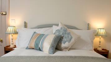 Una cama bien hecha con ropa blanca y varios almohadones, dos lámparas de noche y una mesita de madera.