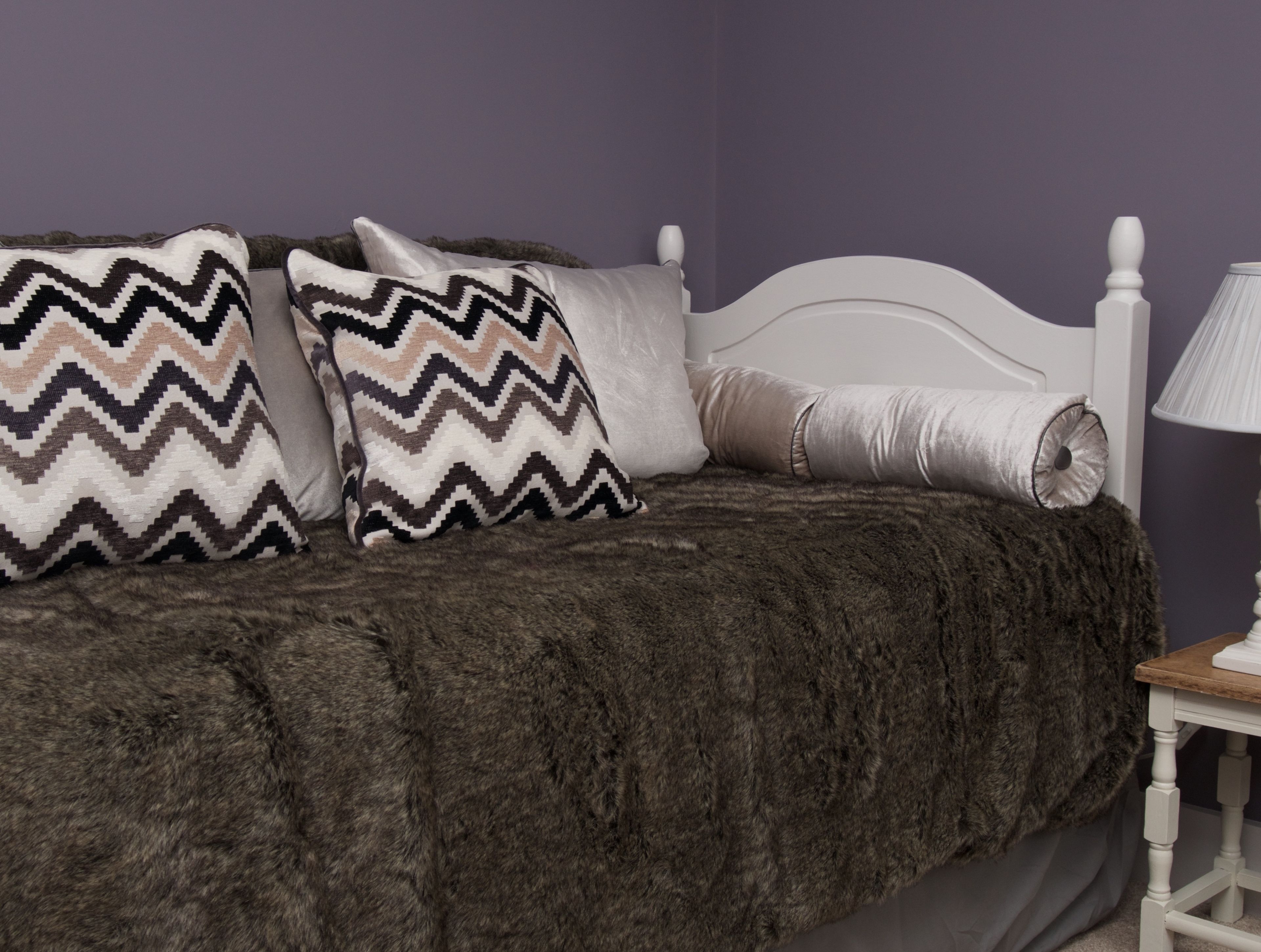 A bed with a dark, fluffy blanket and two patterned pillows, a white headboard, and a bedside table with a lamp.
