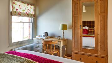 A bedroom with a bed, a wooden desk, a chair, a lamp, a wardrobe, and a window with a floral blind.