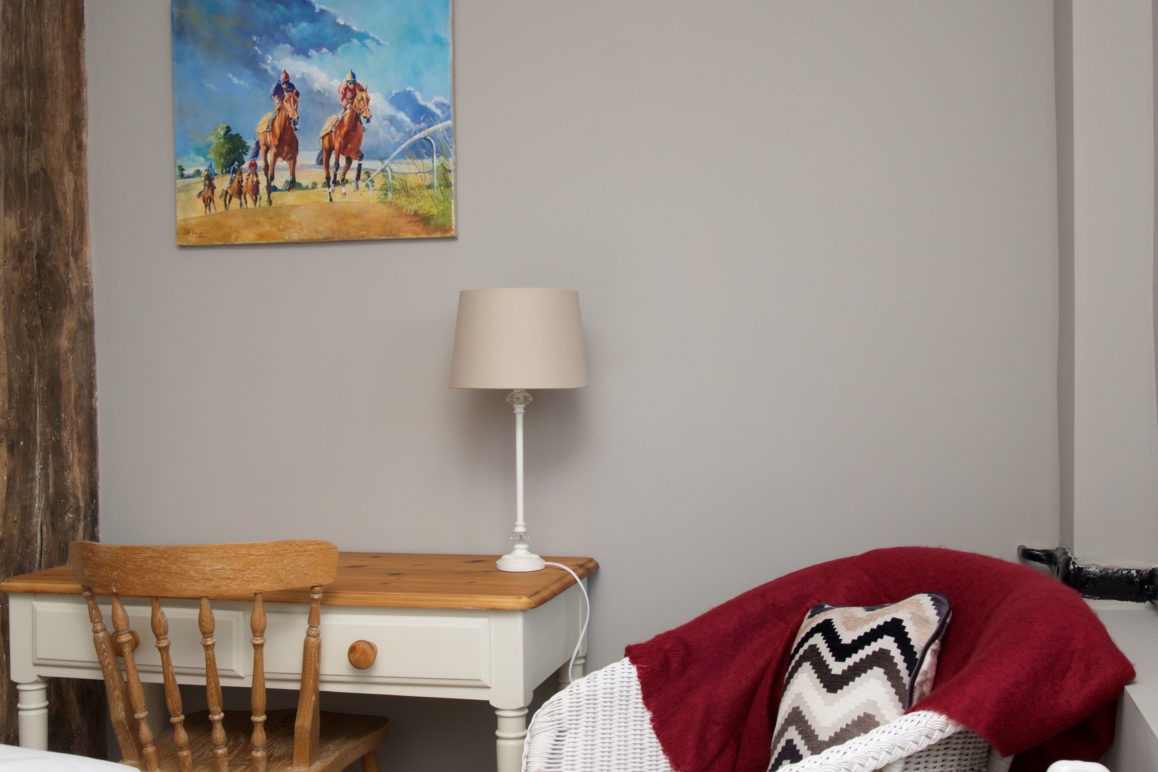 A bedroom with a wooden chair, a white bedside table, a lamp, a red blanket, and a painting of horses on the wall.