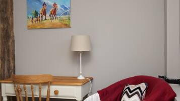 Un dormitorio con una silla de madera, una mesita de noche blanca, una lámpara, una manta roja y un cuadro de caballos en la pared.