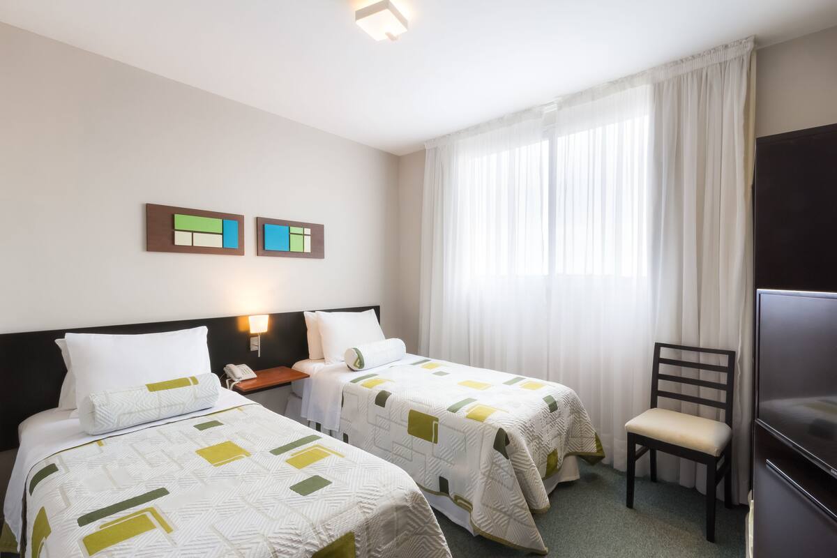 A hotel room with two beds, a TV, a chair, and a nightstand with a lamp.