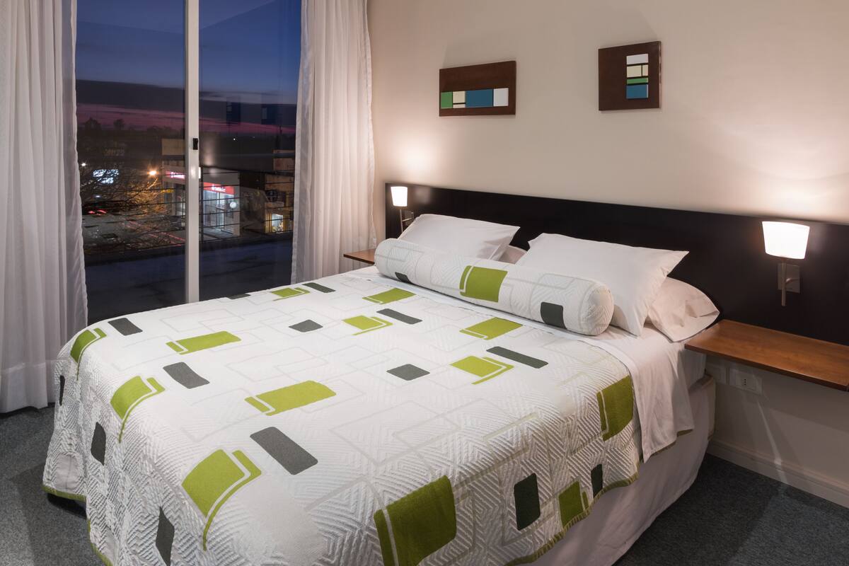 A hotel room with a large bed, two bedside tables with lamps, a view of the city at dusk, and a window with curtains.