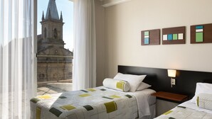 A hotel room with two beds, a nightstand, a wall-mounted lamp, and a view of a church through sheer curtains. - Gran Hotel Brisas del Hum (Mercedes)