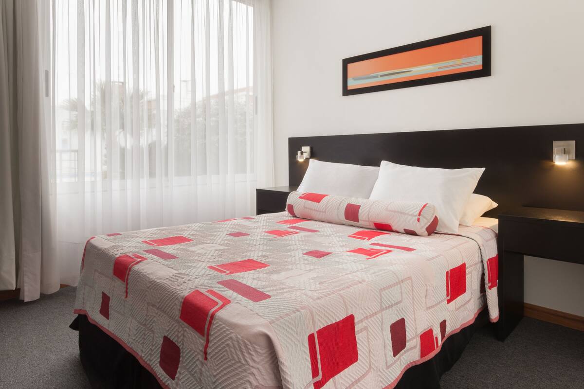 A hotel room with a bed, a nightstand, a wall-mounted lamp, and a window with sheer curtains.