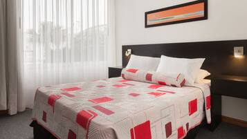 A hotel room with a bed, a nightstand, a wall-mounted lamp, and a window with sheer curtains.
