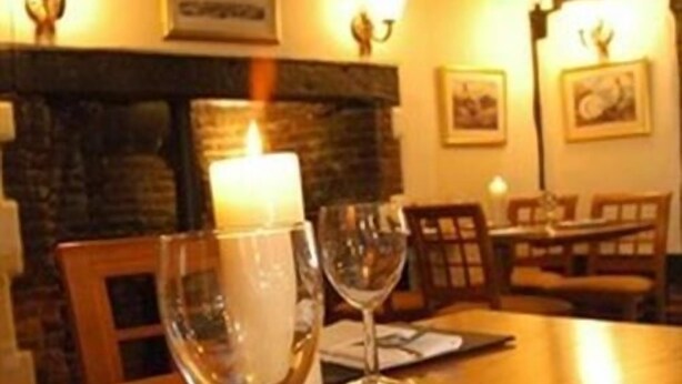 A dining area with a wooden table, two wine glasses, a lit candle, and a stone fireplace in the background. - The New Inn - Kidmore End (Reading)