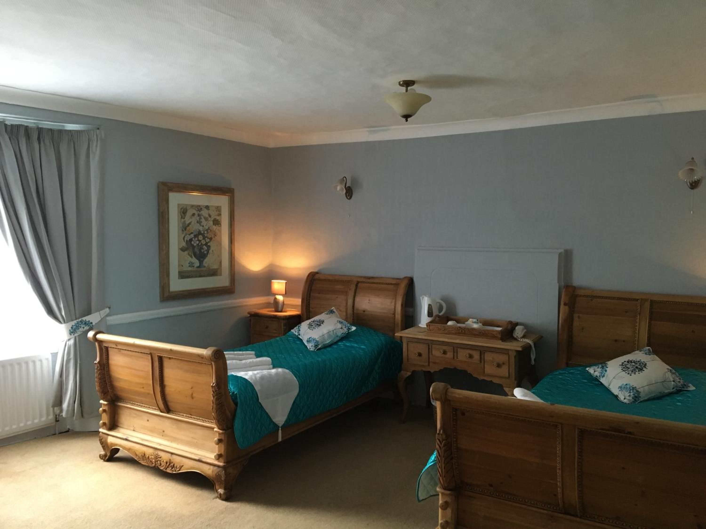 Une chambre avec deux lits en bois, une table de chevet avec une lampe, un tableau encadré au mur et un ventilateur de plafond.