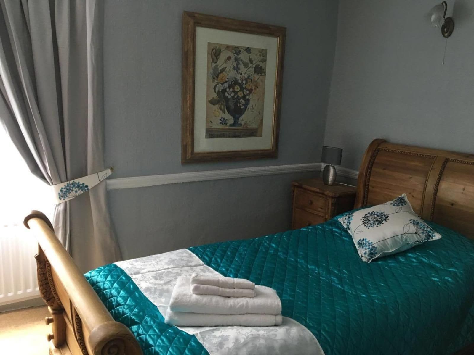 A bedroom with a bed, a nightstand, a lamp, a framed picture on the wall, and a window with curtains.