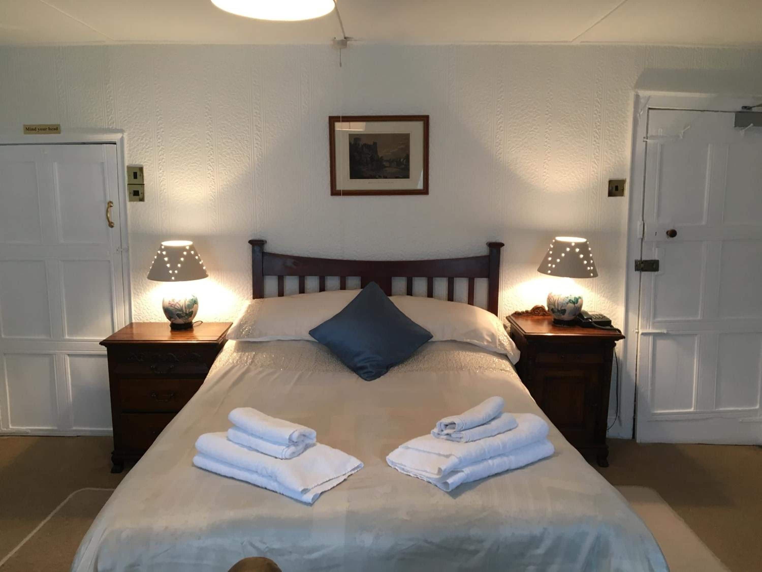 A neatly made bed with folded towels, two bedside lamps, a framed picture on the wall, and a door on the left.