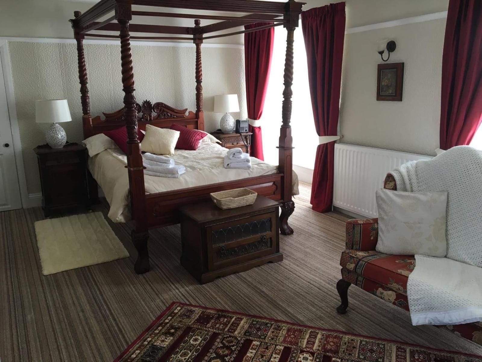 A four-poster bed with a wooden frame, a bedside table with a lamp, a red upholstered chair, and a patterned rug on the floor.