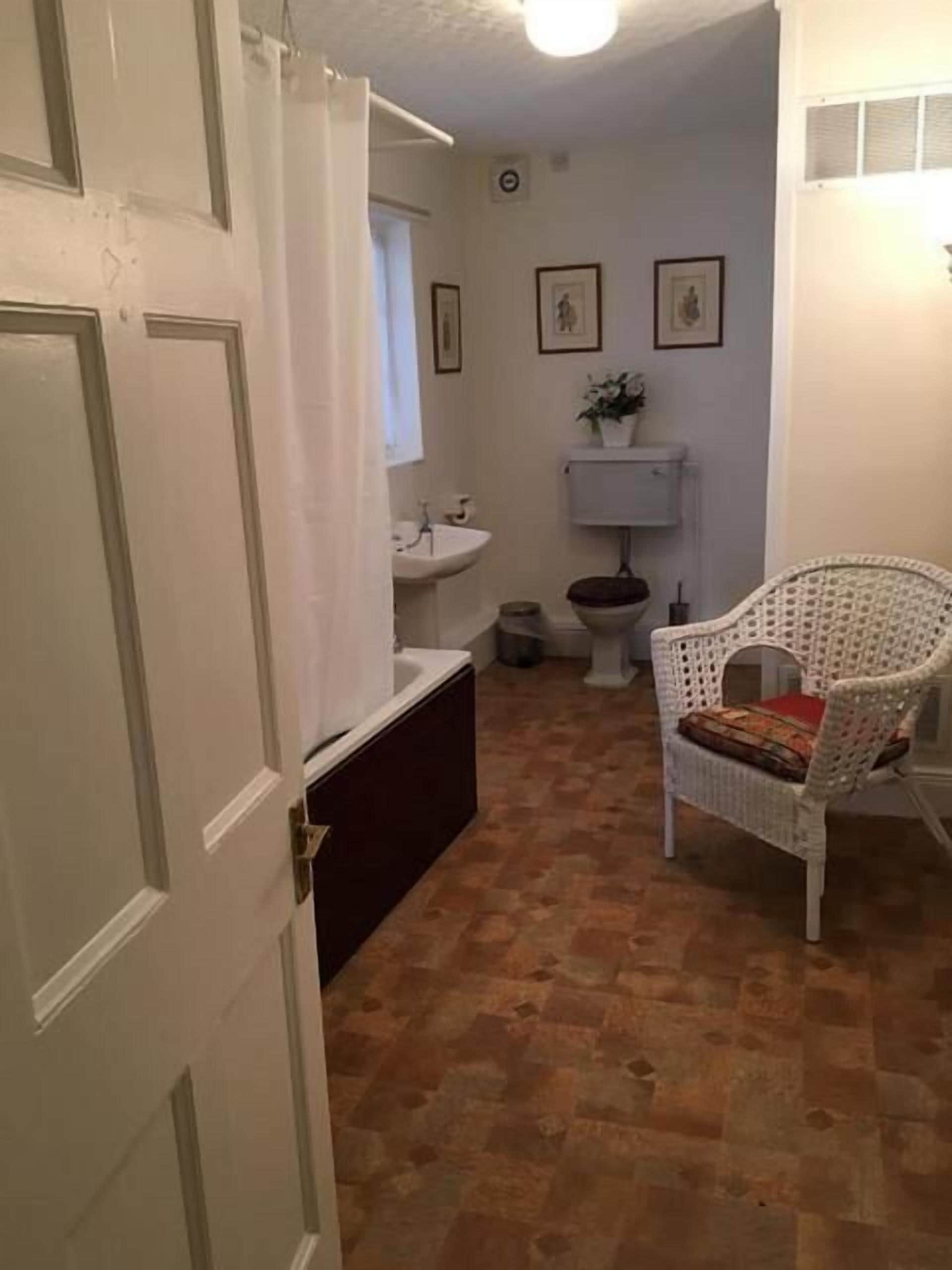Ein Badezimmer mit einer weißen Badewanne, einem Waschbecken und einer Toilette. Ein Korbstuhl mit Kissen steht bereit. Der Boden ist mit Fliesen ausgelegt.