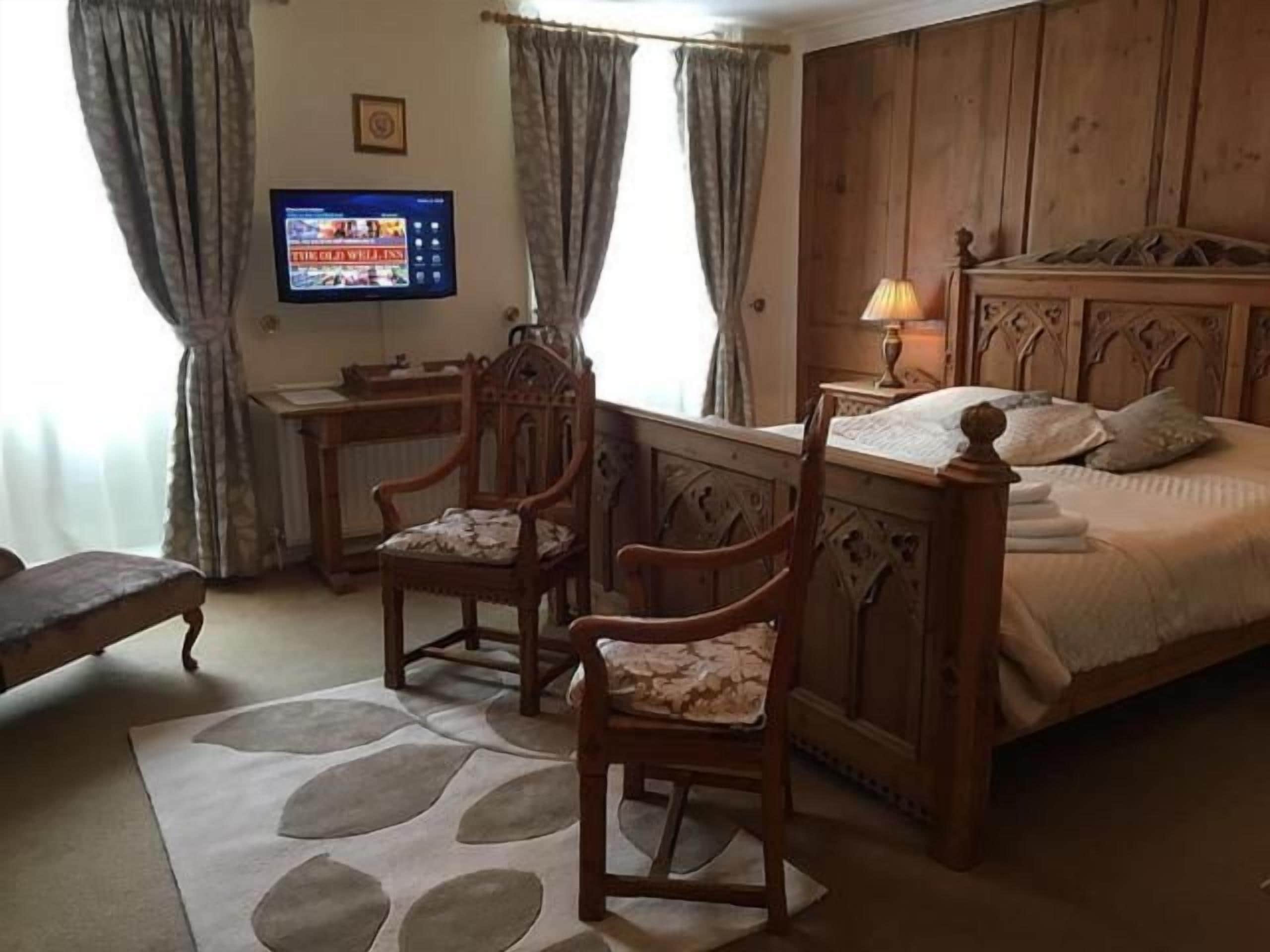 Une chambre à coucher avec un lit en bois, deux chaises, un bureau, une télévision et une fenêtre avec des rideaux.