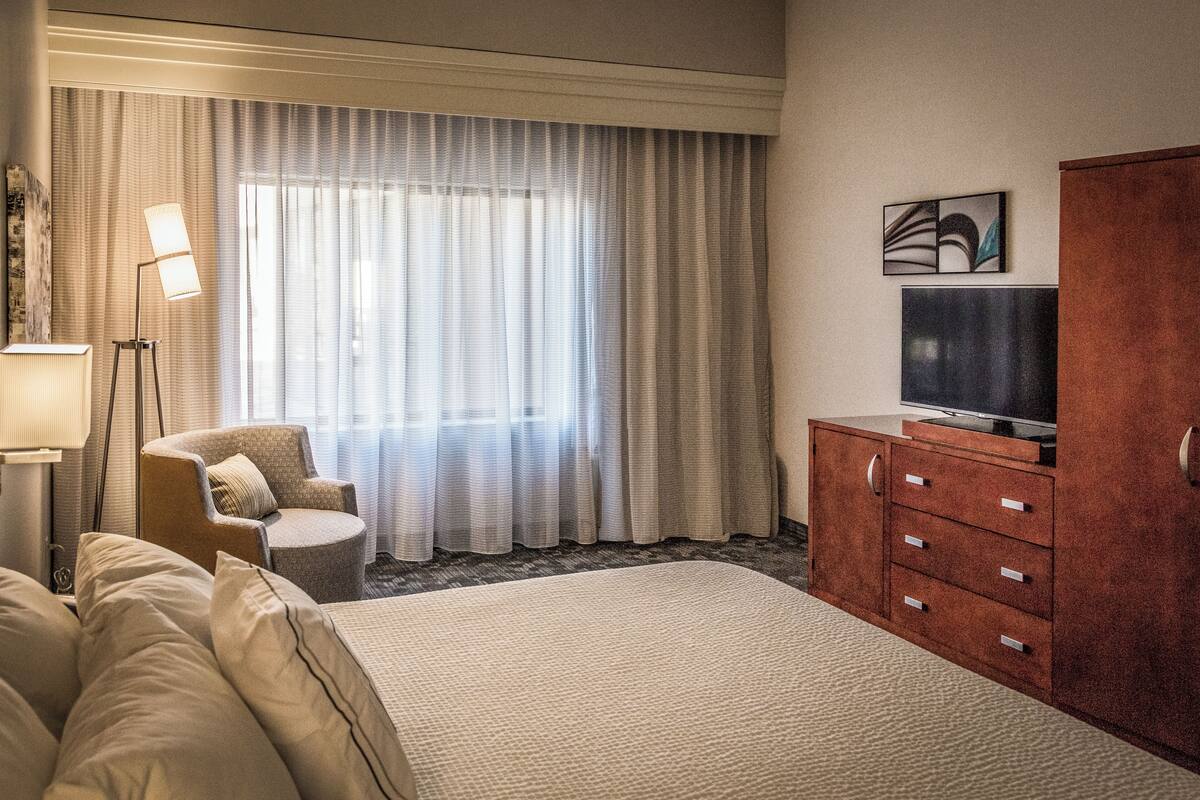A hotel room with a bed, a chair, a desk, a TV on a cabinet, and a window with curtains.