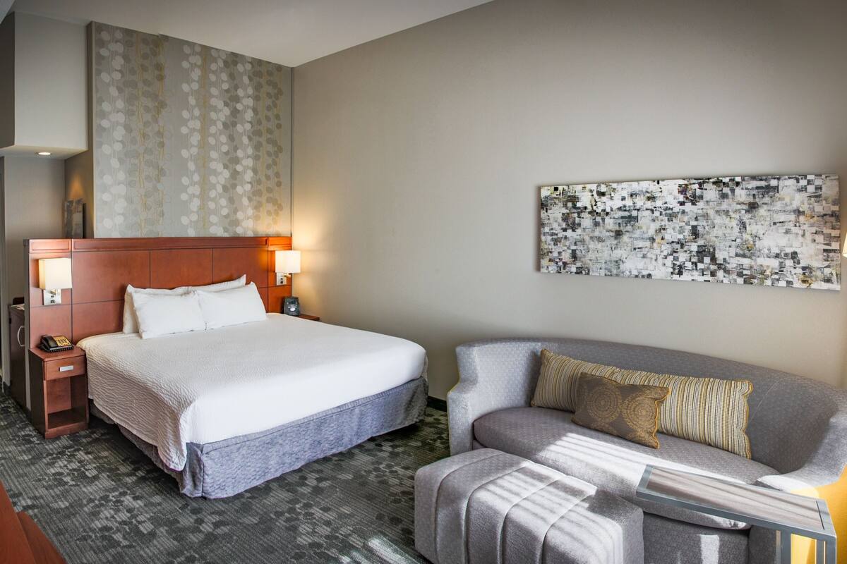 A hotel room with a bed, a sofa, a wall art piece, a lamp, and a bedside table.