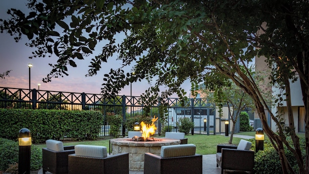 An outdoor seating area with wicker furniture and a central fire feature, surrounded by greenery and a fence.