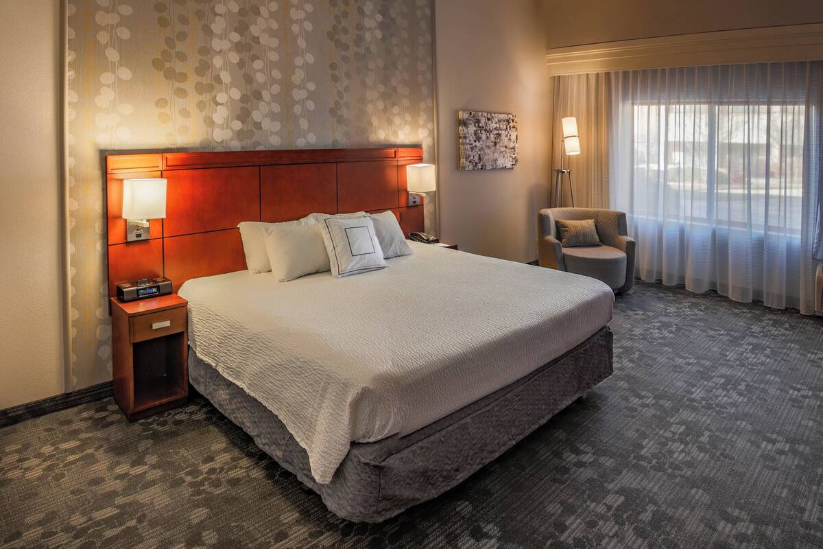 A hotel room with a large bed, two bedside lamps, a nightstand, a chair, and a view of the outdoors through sheer curtains.