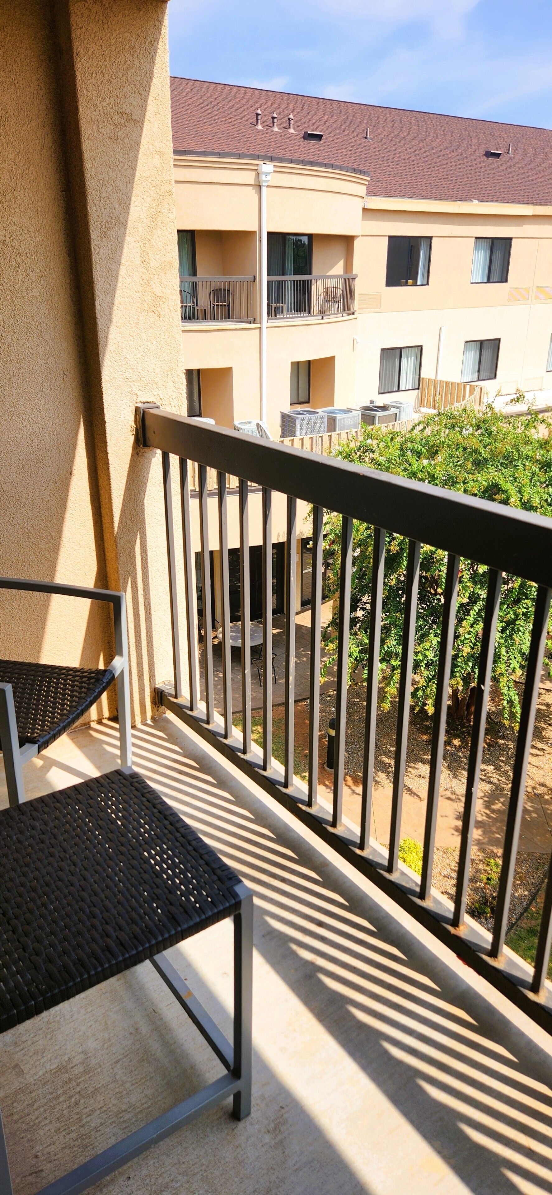 Uma varanda com guarda-corrimão de metal, uma cadeira e uma mesa. Ao fundo, um edifício com várias janelas e varandas.