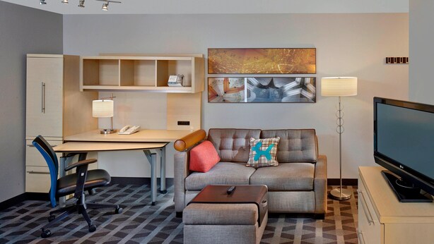 A modern hotel room with a sofa, ottoman, desk, chair, TV, lamp, and artwork.