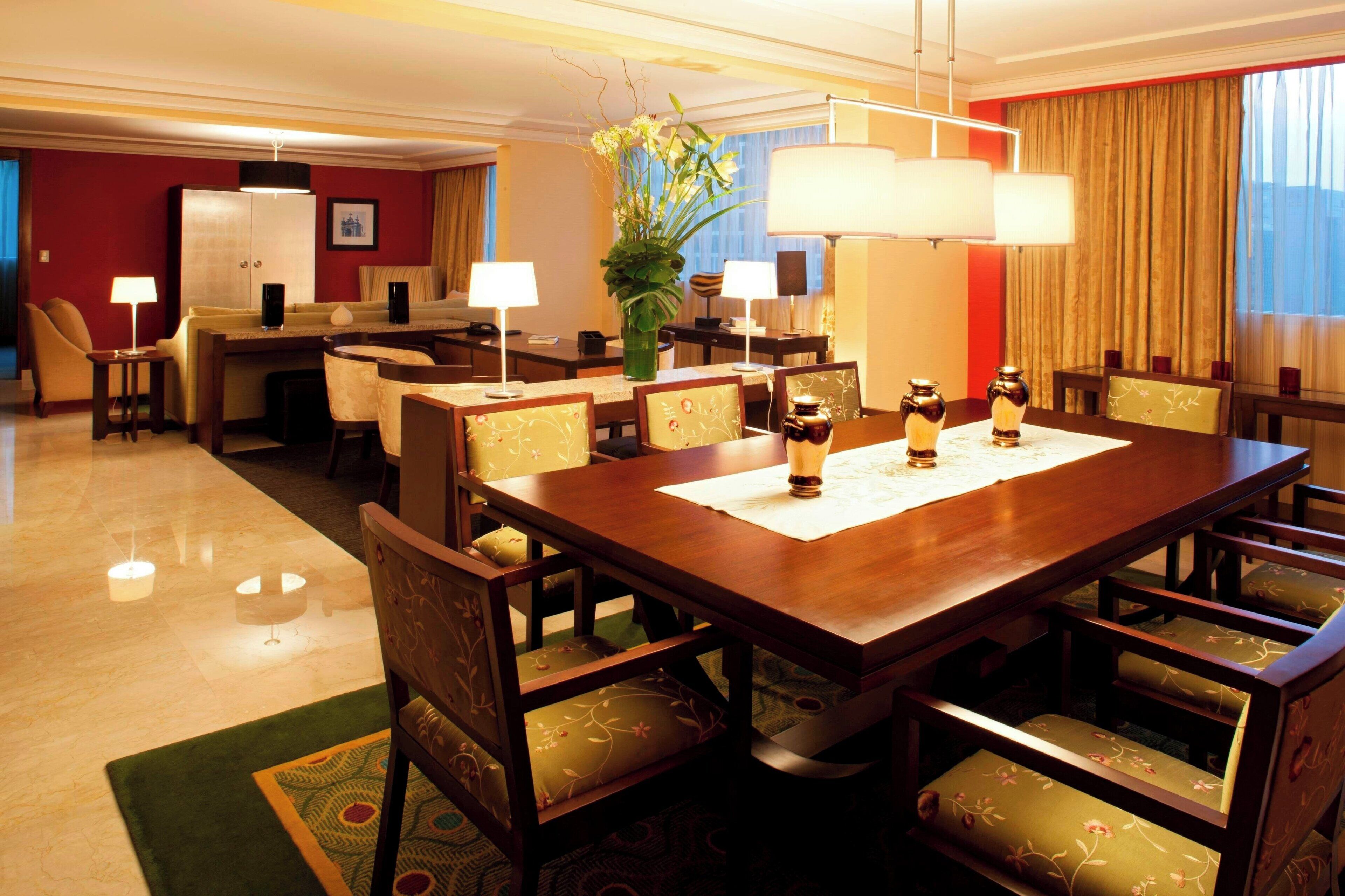 Une salle à manger d’hôtel avec une grande table en bois, plusieurs chaises, un lustre et une plante en pot.