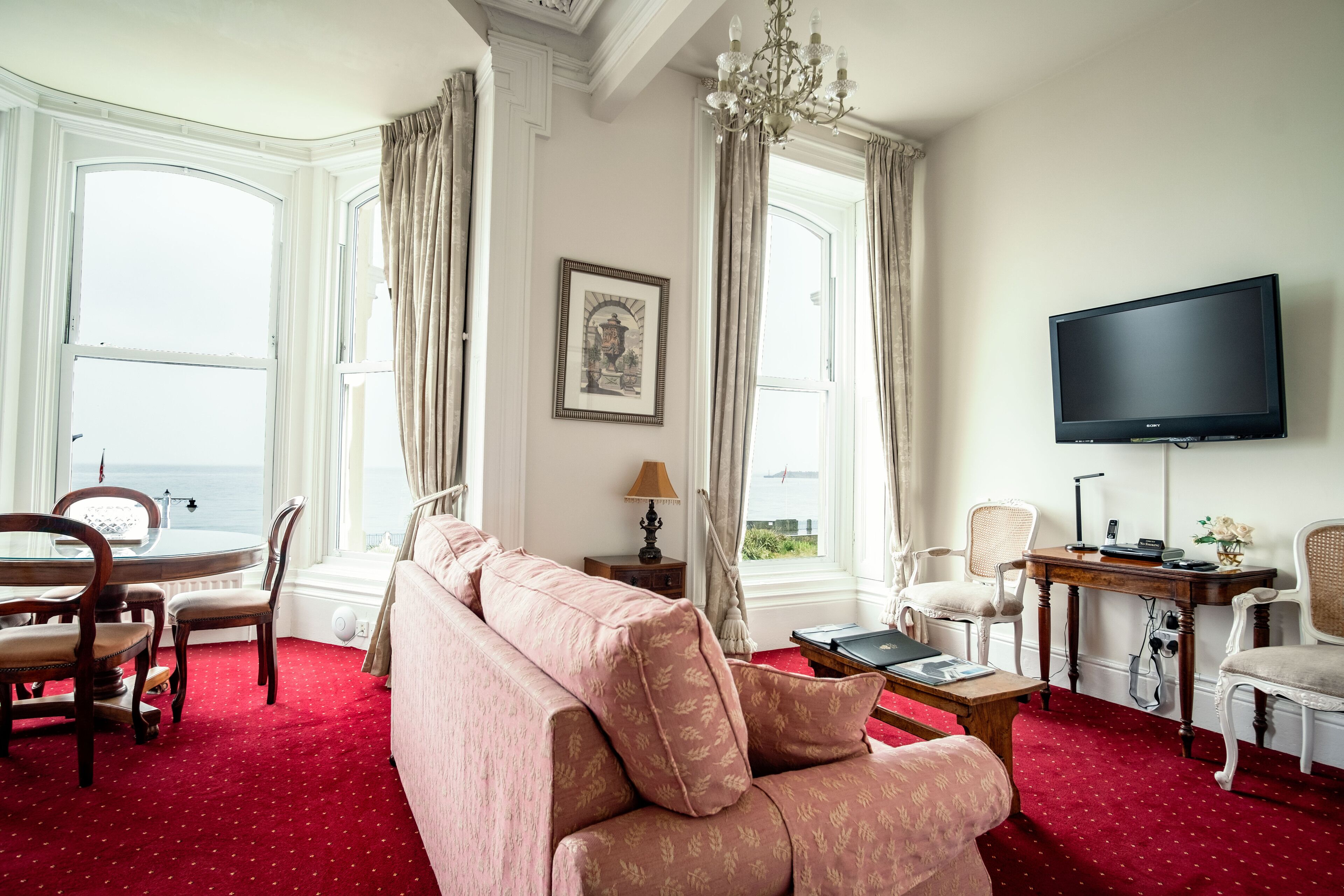 A room with a red carpet, a sofa, a TV, a desk, and a view of the sea.