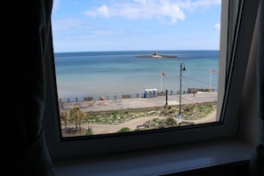 Vista desde una ventana con vista a una playa, una pequeña isla, una zona arenosa con una valla y algunas personas.
