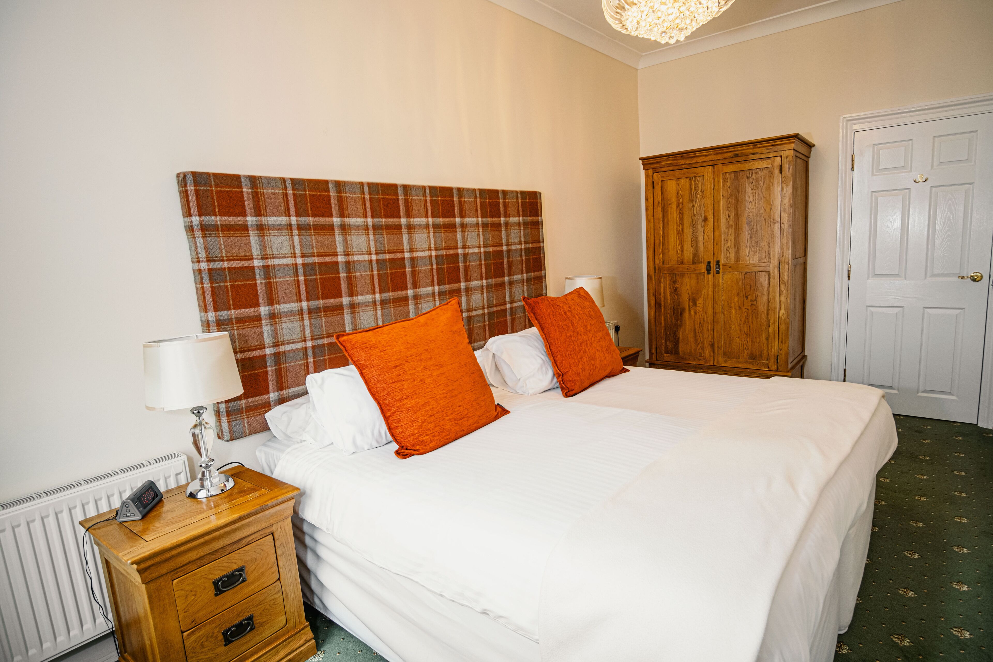 Cama bem arrumada com roupa de cama branca e duas almofadas laranja, mesa de cabeceira de madeira com abajur e cabeceira com padrão xadrez.
