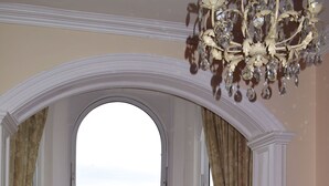 A dining area with a table and chairs, a chandelier, and a view of the sea through an arched window.
