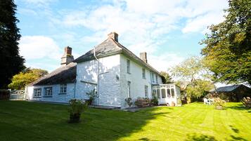 A white stone house with a large lawn, a wooden deck, and a garden area.