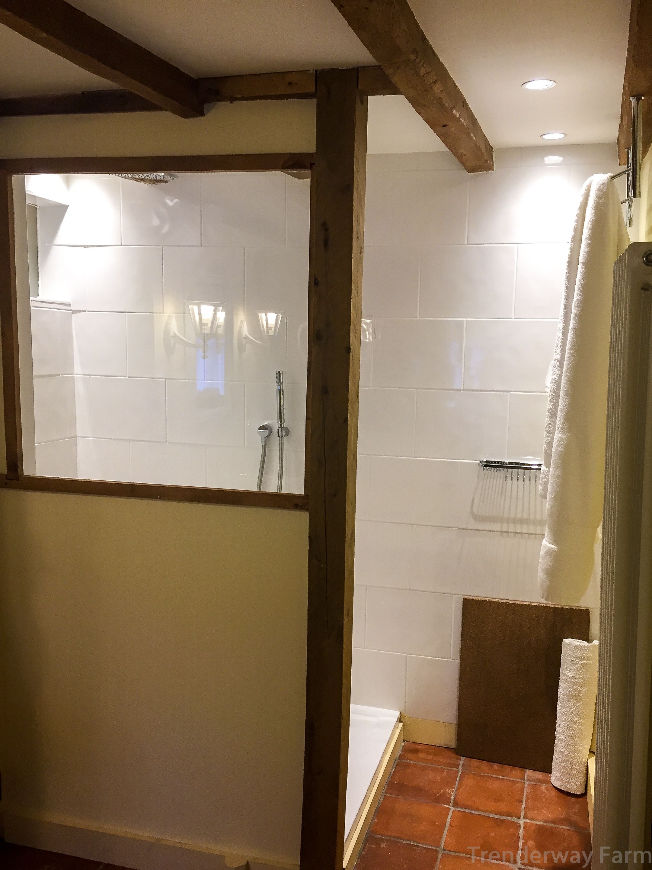 Un baño con paredes de azulejos blancos, techo de vigas de madera, zona de ducha y un perchero para toallas.