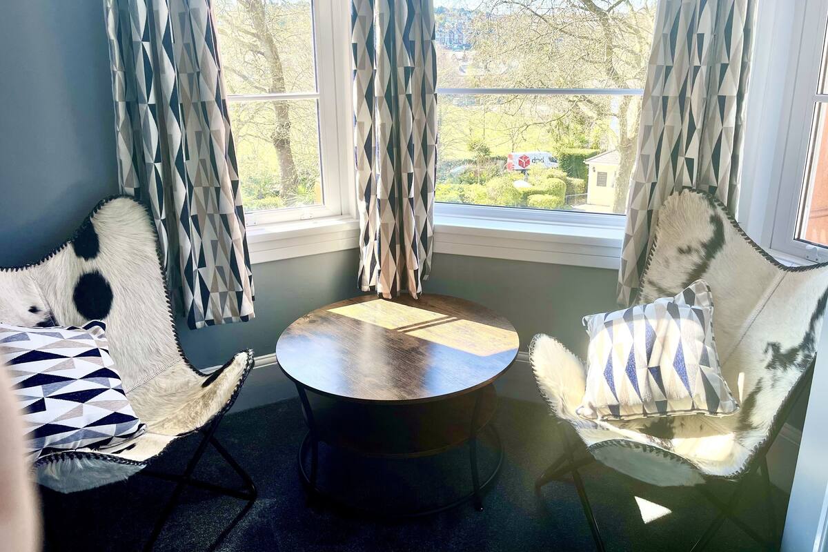 A corner seating area with two chairs and a small table, a window with curtains, and a view of greenery outside.